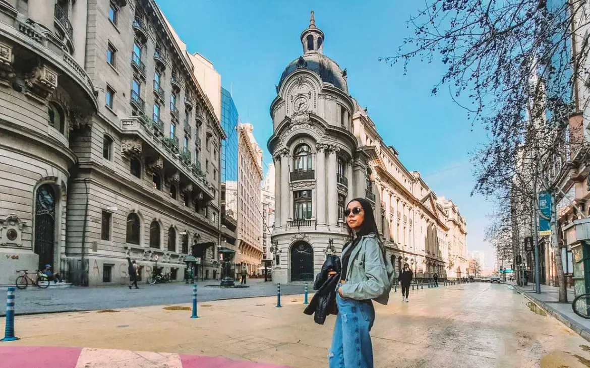 Uma mulher de óculos escuros está posando em uma rua ampla e ensolarada, cercada por edifícios históricos ornamentados, no centro de Santiago do Chile, um ótimo local para considerar quando pensar em onde ficar em Santiago.