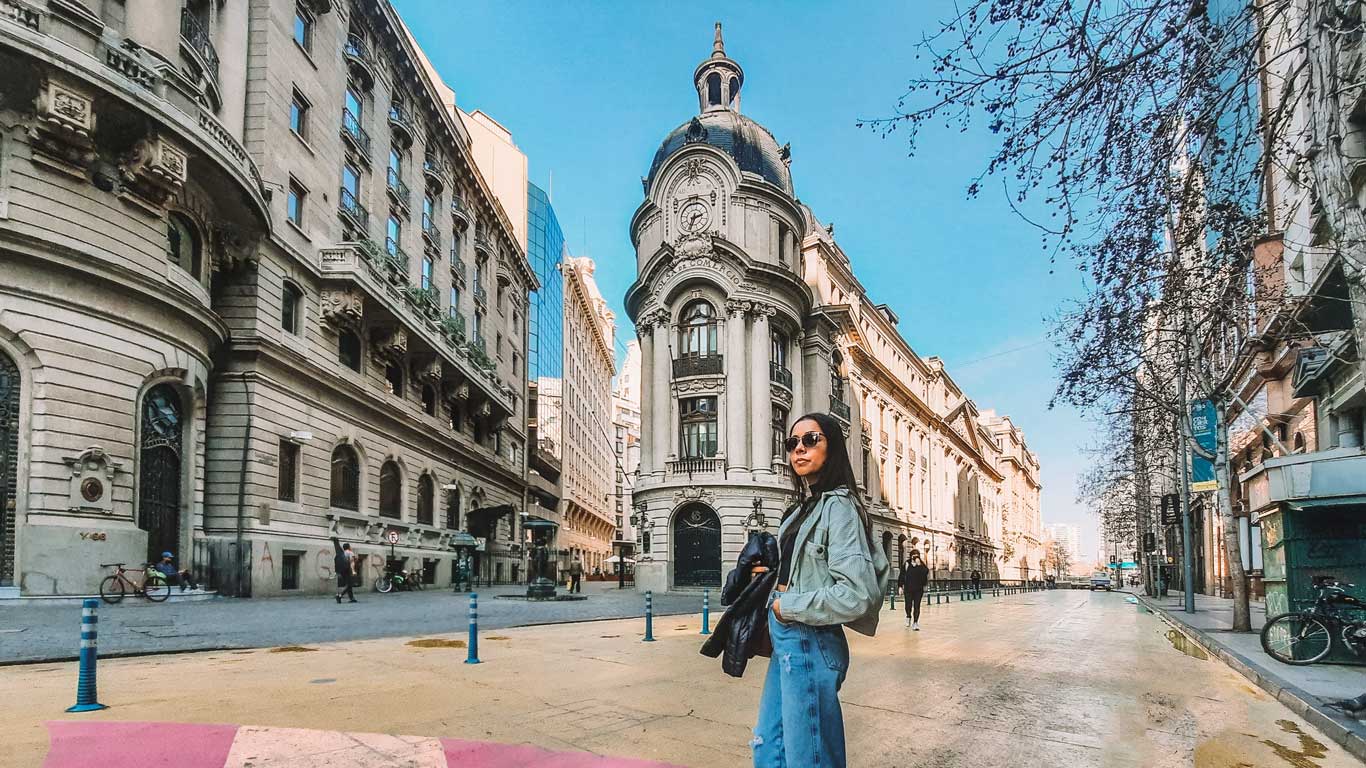 Uma mulher de óculos escuros está posando em uma rua ampla e ensolarada, cercada por edifícios históricos ornamentados, no centro de Santiago do Chile, um ótimo local para considerar quando pensar em onde ficar em Santiago.