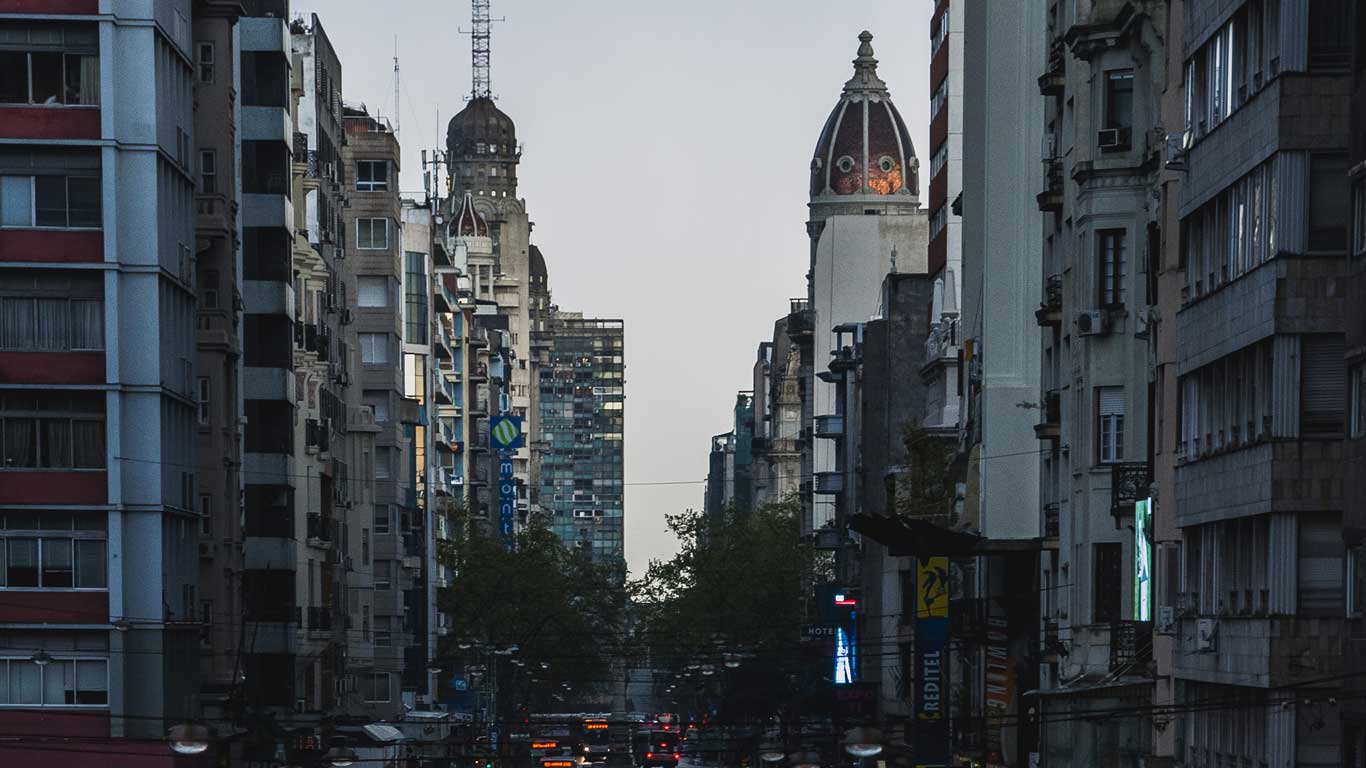 Vista da Avenida 18 de Julho em Montevidéu ao entardecer, cercada por edifícios altos e históricos. No horizonte, torres arquitetônicas se destacam, enquanto as luzes da cidade começam a brilhar. 
