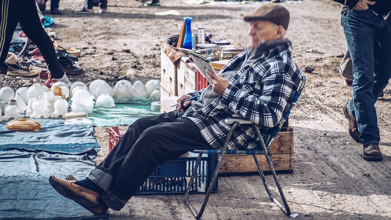 Homem idoso sentado em uma cadeira na Feira Tristán Narvaja em Montevidéu, lendo um jornal. Ao redor, vários itens usados estão à venda, incluindo luminárias e objetos de vidro. A feira é conhecida por sua diversidade de produtos e atmosfera acolhedora.