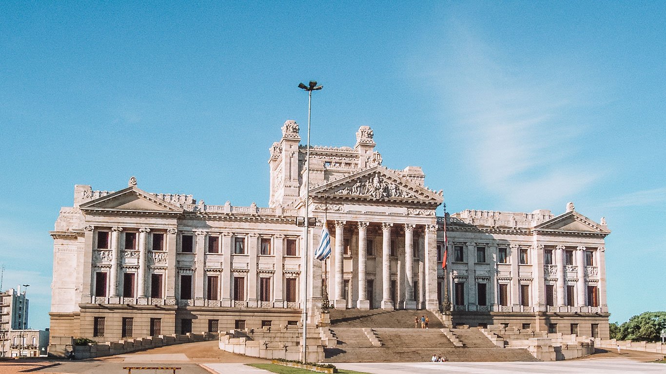Fachada do Palácio Legislativo de Montevidéu, um imponente edifício neoclássico com colunas e esculturas detalhadas. A bandeira do Uruguai está hasteada na frente do palácio, que é um importante símbolo político e histórico do país.