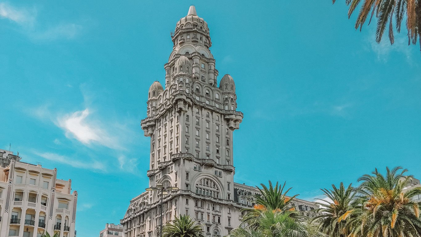 Vista do Palácio Savo durante o dia sob um céu azul claro. Este icônico edifício, localizado em Montevidéu, Uruguai, destaca-se por sua arquitetura neorrenascentista com detalhes ornamentais e uma torre proeminente. Palmeiras se elevam em primeiro plano, adicionando um toque tropical ao ambiente urbano.