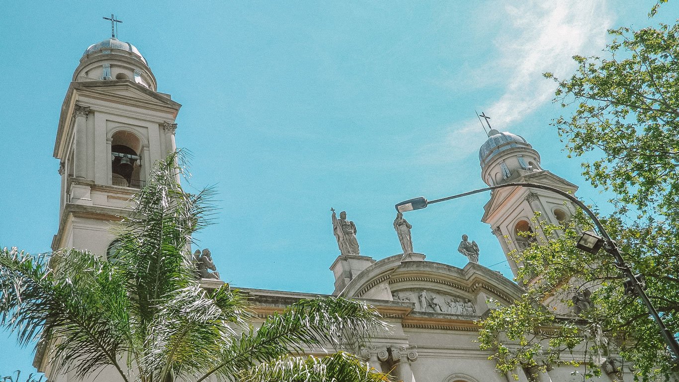 Fachada da Catedral Metropolitana de Montevidéu, com suas torres gêmeas e estátuas decorativas no topo. A catedral é cercada por palmeiras e árvores, sob um céu azul claro.