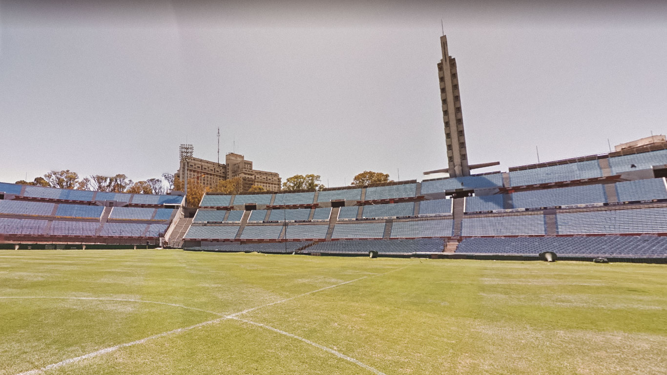 Vista interna do Estádio Centenário em Montevidéu, mostrando a arquibancada com assentos azuis e o gramado verde bem cuidado. Ao fundo, a Torre de los Homenajes se destaca como um símbolo icônico do estádio.