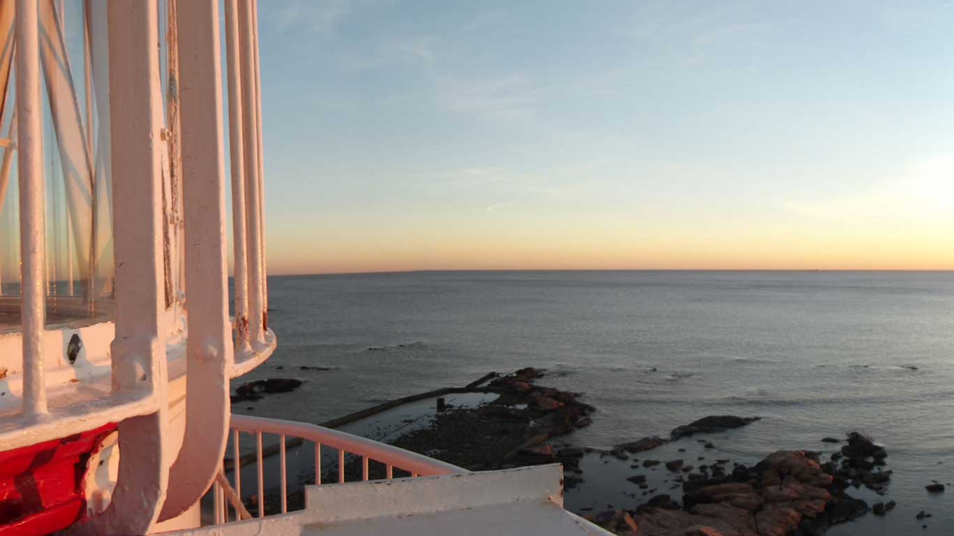 Vista do Farol de Punta Carretas em Montevidéu ao entardecer, com a estrutura branca do farol em primeiro plano e o mar calmo ao fundo. O céu está tingido de tons de laranja e azul, criando uma atmosfera serena e pitoresca.