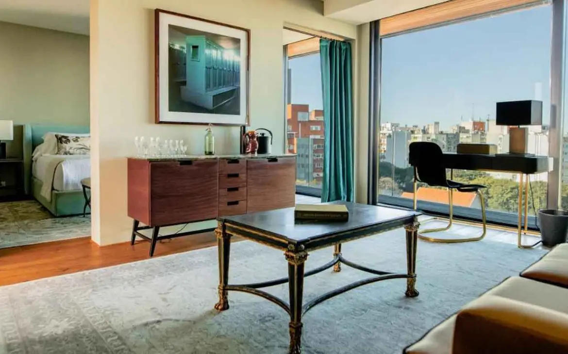 A imagem mostra uma elegante suíte de hotel com uma decoração moderna e vista panorâmica da cidade de Montevidéu. O quarto apresenta uma cama confortável, uma mesa de escritório e uma área de estar bem iluminada com grandes janelas que oferecem uma vista impressionante dos edifícios ao redor.