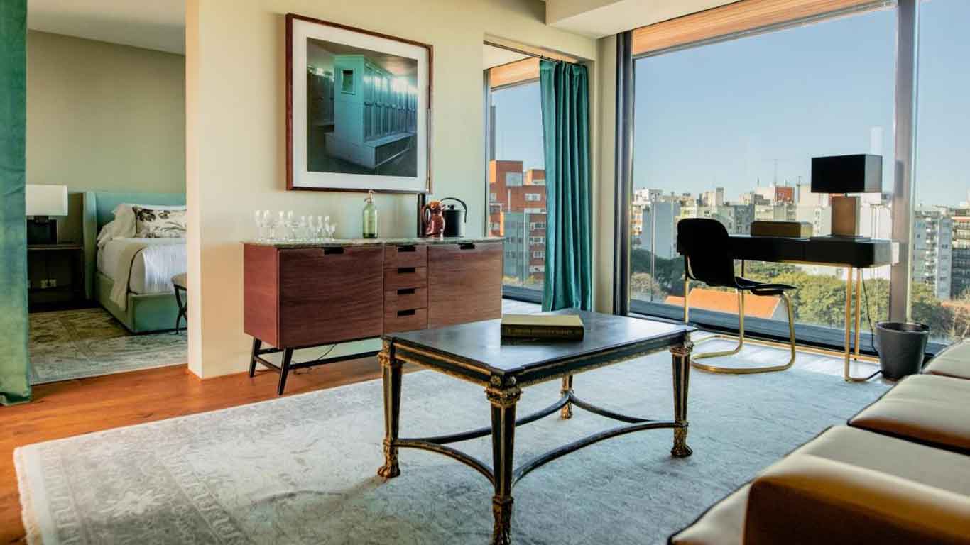 A imagem mostra uma elegante suíte de hotel com uma decoração moderna e vista panorâmica da cidade de Montevidéu. O quarto apresenta uma cama confortável, uma mesa de escritório e uma área de estar bem iluminada com grandes janelas que oferecem uma vista impressionante dos edifícios ao redor.