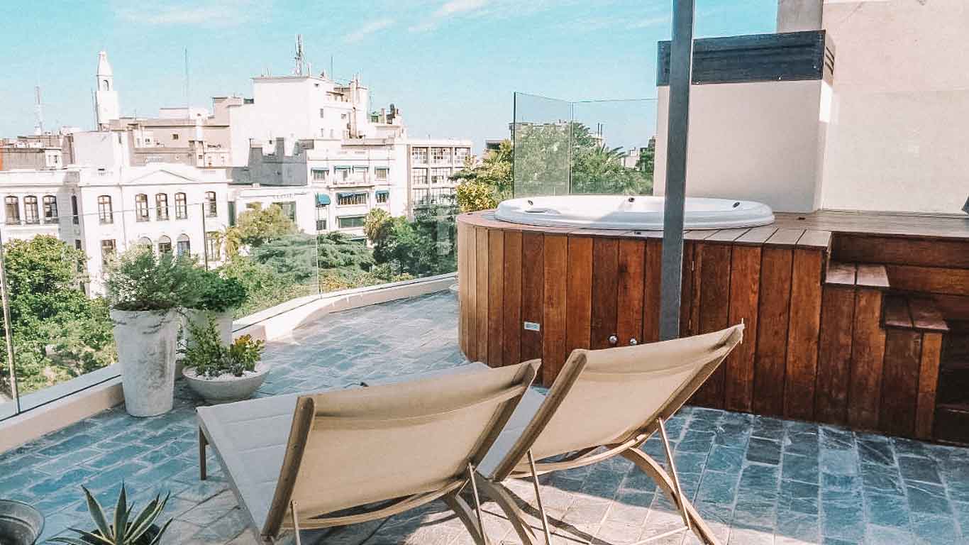 Imagem de um terraço do Alma Histórica Boutique Hotel Montevidéu, mostrando duas espreguiçadeiras voltadas para uma vista panorâmica da cidade, com prédios históricos ao fundo e uma jacuzzi de madeira ao lado.