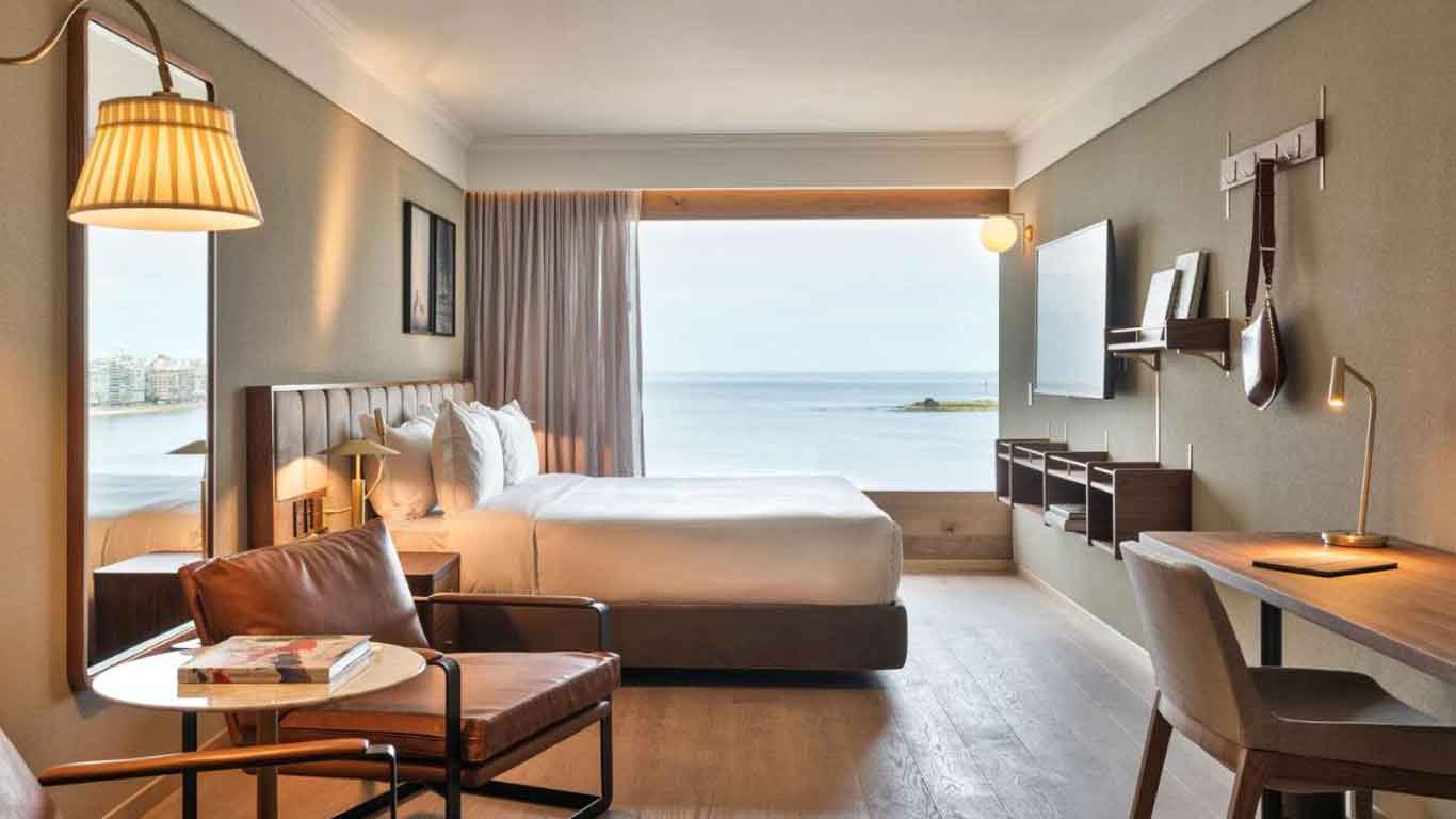 Quarto do Hotel Costanero MGallery em Montevidéu, apresentando uma cama de casal com vista para o mar através de uma ampla janela, uma cadeira confortável com uma mesa de centro e um ambiente de decoração moderna e aconchegante.