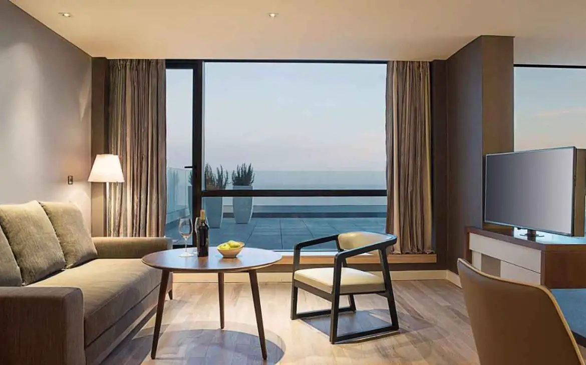 A imagem mostra uma sala de estar moderna e aconchegante em um quarto de hotel, com móveis elegantes e uma grande janela que oferece uma vista panorâmica para o mar. A sala é equipada com um sofá, uma mesa de centro com uma garrafa de vinho e copos, e uma televisão. Este é um exemplo de acomodação de luxo no Hyatt Hotel em Montevidéu.