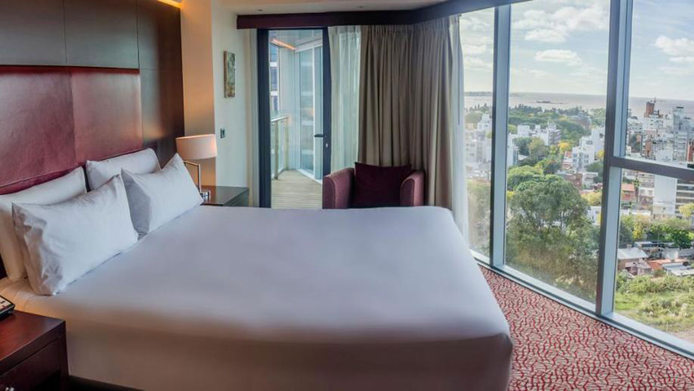 Quarto do Hilton Garden Inn Montevideo, apresentando uma cama de casal grande com lençóis brancos, uma poltrona roxa próxima à janela e uma vista panorâmica da cidade e do rio. A decoração é moderna, com janelas amplas que permitem entrada de muita luz natural.