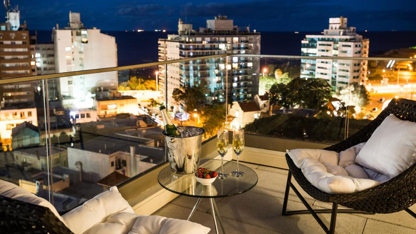 Varanda do hotel Own Montevideo, à noite, com vista para a cidade iluminada e o mar ao fundo. A varanda possui duas cadeiras acolchoadas ao redor de uma mesa de vidro com uma champanheira, duas taças de champanhe e um recipiente com morangos.