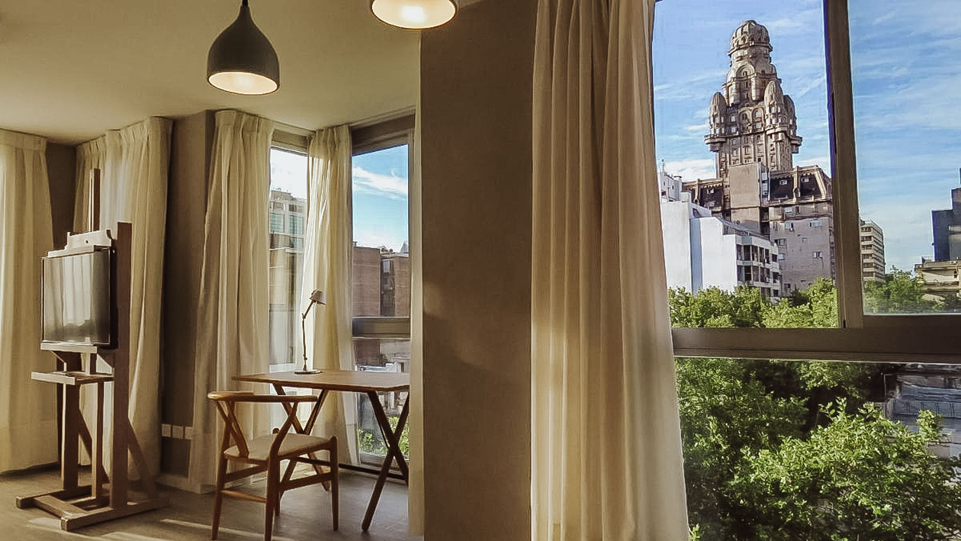 Quarto do Smart Hotel Montevideo, com uma mesa de trabalho e uma cadeira próxima a uma ampla janela com vista para o famoso Palácio Salvo. A decoração é moderna e minimalista, com cortinas claras que permitem a entrada de luz natural.