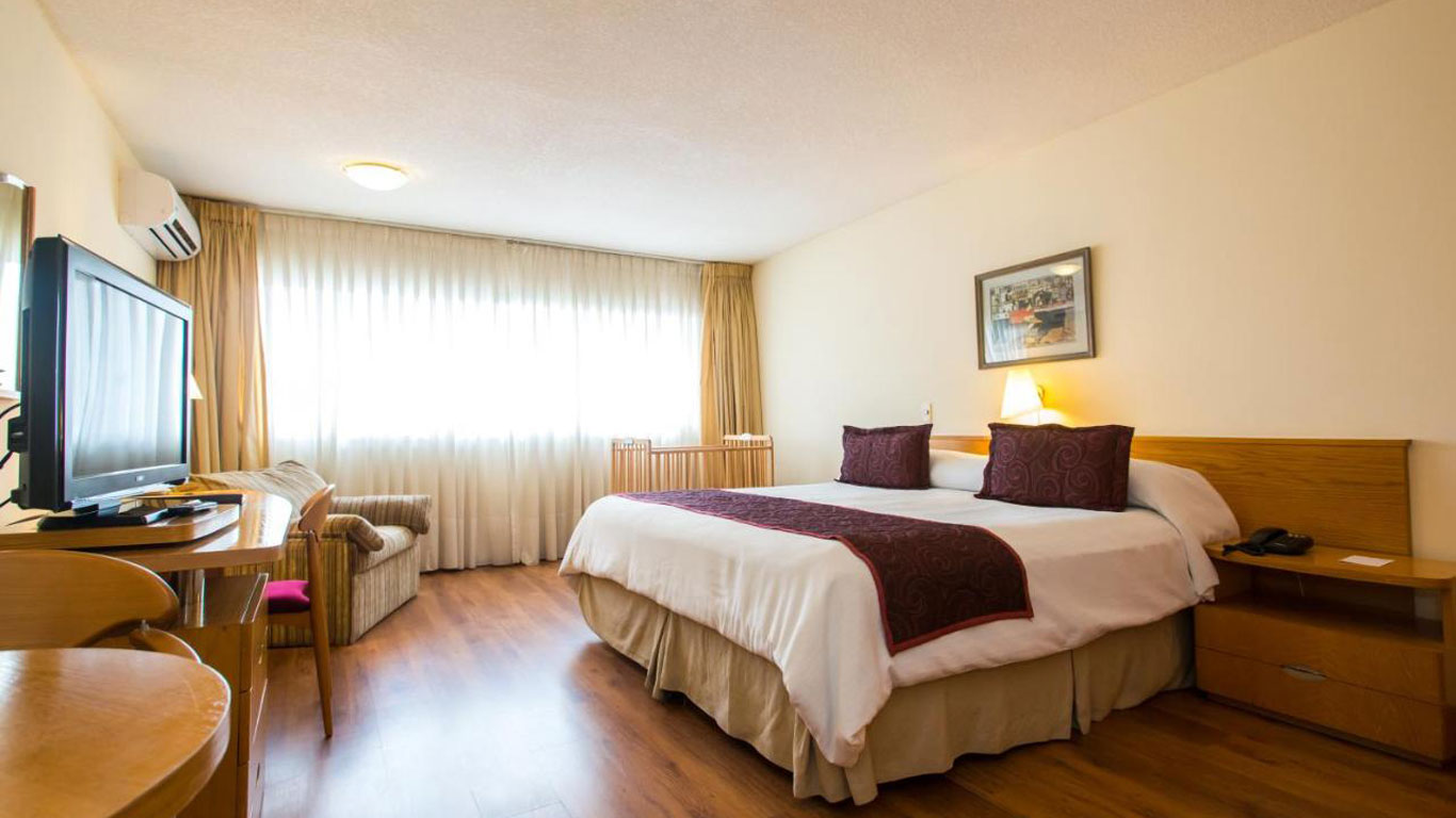 Quarto do Armon Suites Hotel em Montevidéu, apresentando uma cama de casal grande com roupa de cama branca e detalhes em roxo, uma televisão em frente à cama, e uma poltrona ao lado de uma janela ampla com cortinas claras. A decoração é acolhedora e funcional, com piso de madeira e móveis simples.