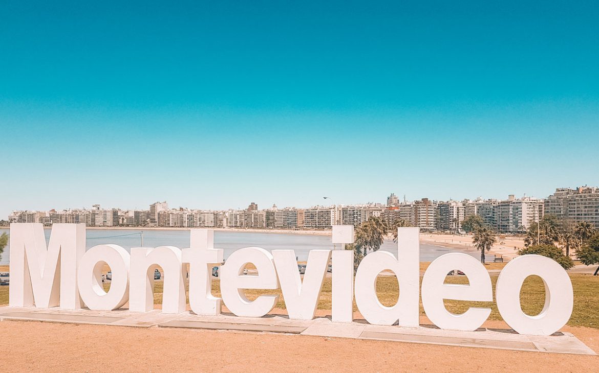 A imagem exibe um grande letreiro escrito "Montevideo" em letras brancas volumosas, posicionado em um parque com uma vista panorâmica da linha do horizonte urbana da cidade ao fundo. O céu é claro e azul, contrastando com o verde da grama e a areia da praia visíveis abaixo do letreiro. Esta foto captura um ponto turístico popular em Montevidéu, ideal para quem deseja imortalizar sua visita com uma foto memorável da capital uruguaia.