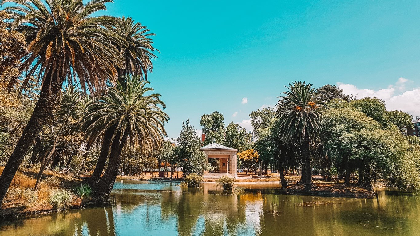 Vista do Parque Rodó em Montevidéu, destacando um lago cercado por palmeiras exuberantes e vegetação diversa. No centro da imagem, há uma pequena construção em estilo clássico à beira da água.