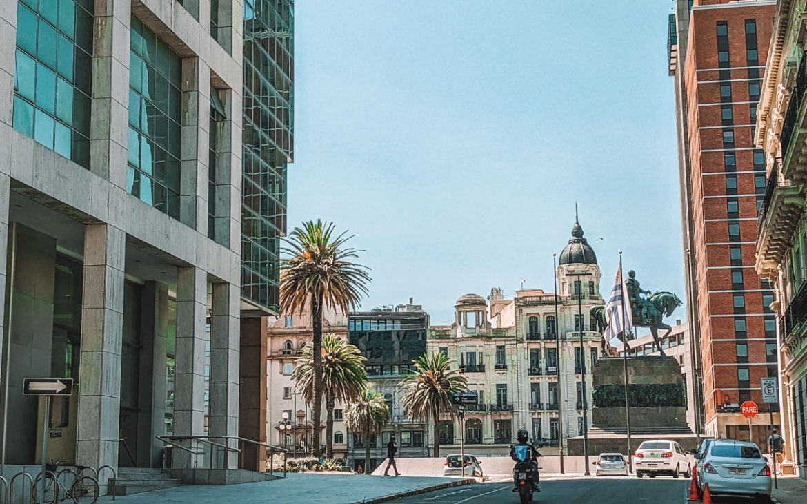 Rua em Montevidéu, destacando um prédio moderno à esquerda e construções históricas ao fundo, incluindo uma estátua e bandeiras. Palmas decoram a paisagem urbana, e uma moto transita pela via. A arquitetura combina elementos modernos e tradicionais, simbolizando a diversidade da cidade.