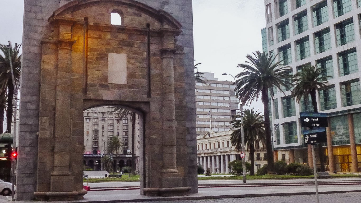 A Puerta de la Ciudadela em Montevidéu, um arco de pedra histórica que marca a entrada para a cidade antiga. Ao fundo, vê-se edifícios modernos e palmeiras, contrastando com a estrutura histórica da porta.