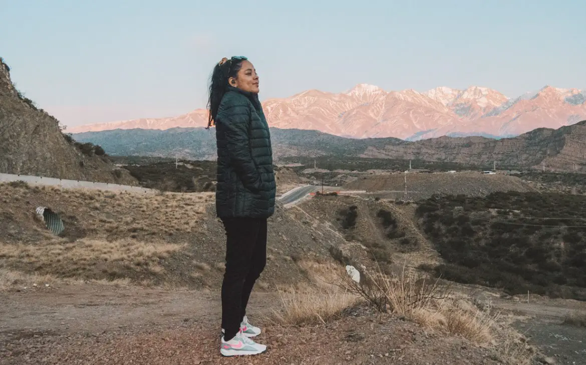 Uma mulher de pé em uma colina em Mendoza, com a Cordilheira dos Andes ao fundo. Ela está usando uma jaqueta preta e tênis brancos, parecendo contente enquanto o sol nasce atrás dela.
