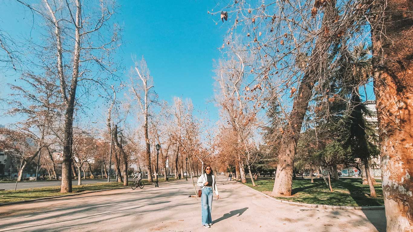 Uma mulher caminha por uma alameda cercada por árvores no bairro Bellas Artes, uma excelente opção de onde ficar em Santiago do Chile, com um céu azul claro ao fundo.