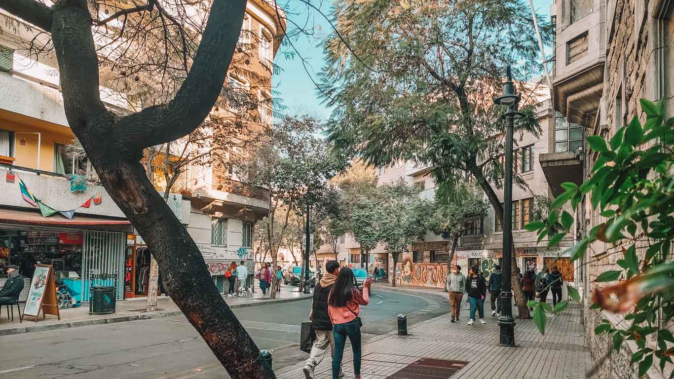 Um casal caminha por uma rua arborizada e movimentada, cercado por lojas e edifícios residenciais no bairro Lastarria, o melhor lugar para ficar em Santiago do Chile.